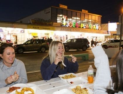 Three happy young women in sweatshirts enjoying fast food takeout across the street from Joe's Playland in Salisbury MA at night