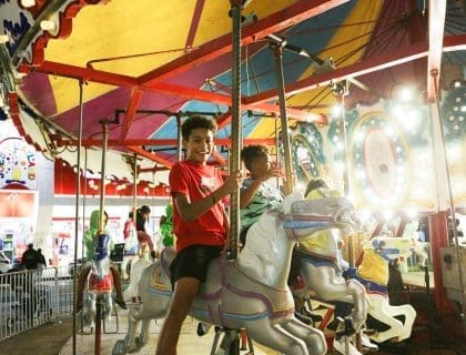 A happy young boy in a red tshirt and black shorts smiling while riding on a children-filled lit-up carousel outdoors at night in Salisbury MA