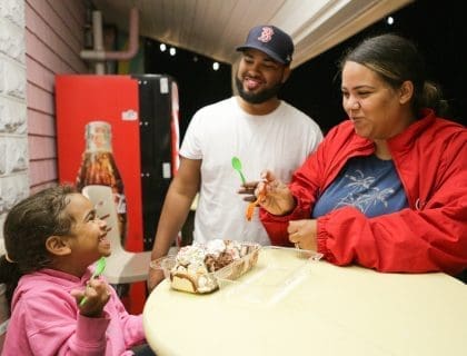 Smiling family including a father in a Boston Red Sox baseball cap, mom in red jacket and navy tee, and young daughter in pink sweatshirt enjoying an ice cream sundae together in front of a coca cola vending machine in Salisbury MA