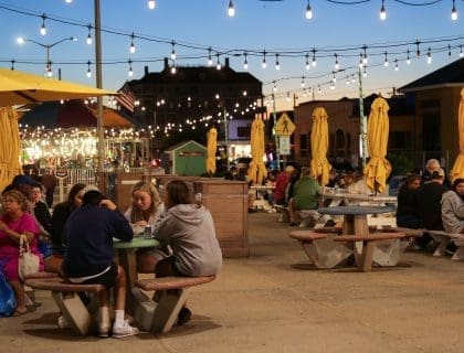 Four Teens sitting around an outdoor table near a lit carousel under string lights with others sitting at tables in the background in Salisbury MA