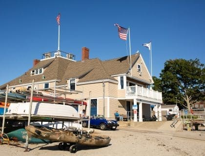 View of a stack of kayaks and a boat lift outside in the sand to the left of the Swampscott Yacht Club located in Fish House on a clear blue sky day