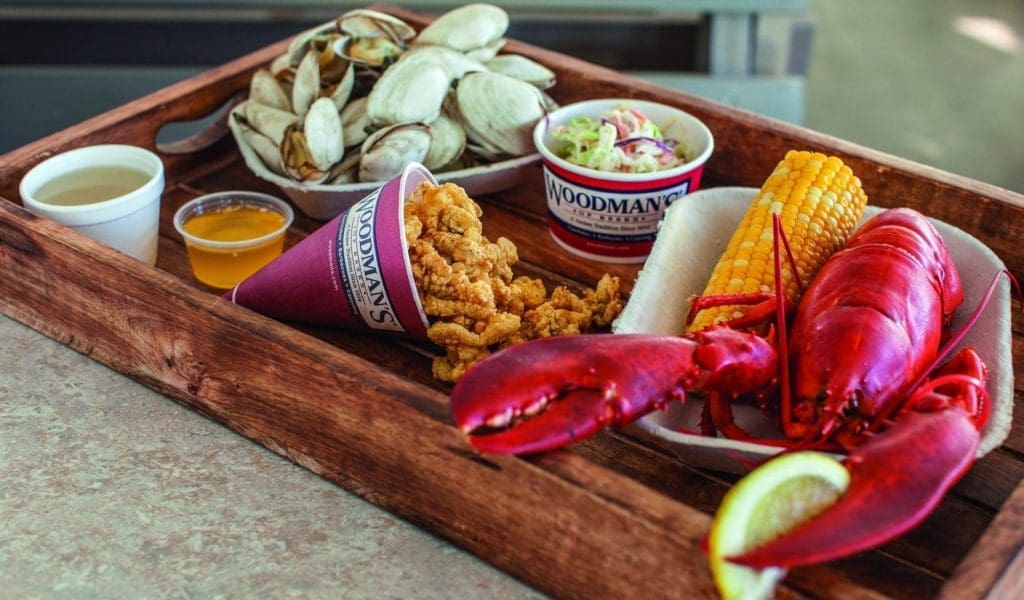 A closeup of a delicious display from right to left of fresh lobster, fried clams, steamed clams, slaw, a small cup of melted butter in a wooden tray from Woodmans in Essex MA