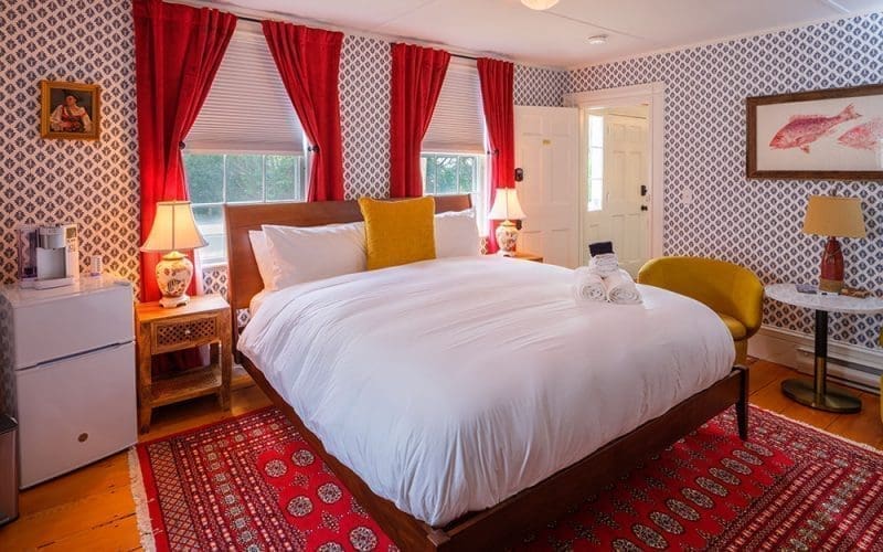 A room with blue & white printed wallpaper with framed pictures on the wall, bright red open curtains on each window, 2 wooden nightstands with an antique looking lamp on each, a mini fridge, a yellow chair, a bed with white bedding and one yellow pillow with clean towels placed upon it, and a printed red carpet underneath the bed at Boutique Inn called the Sally Webster Collection in Rockport MA