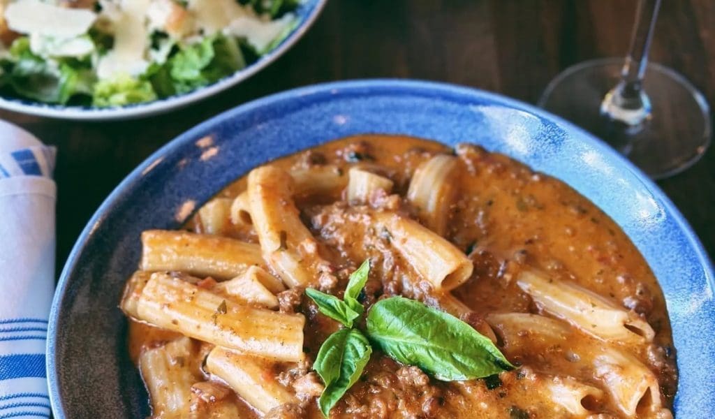 A close-up of rigatoni bolognese with other freshly made Italian food on blue plates and a glass of red wine in the background from Paolo's in Essex MA