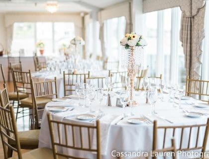 A brightly lit Atlantic Ocean Room at the Elks at Bass Rocks in Gloucester, MA with elegantly set round tables with white and yellow floral centerpieces and white tablecloths with gold chairs.