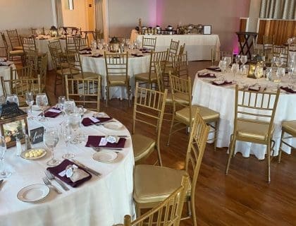 Elegantly set round tables with gold chairs and pink lighting for an event in The Atlantic Ocean Room at the Elks at Bass Rocks in Gloucester, MA.