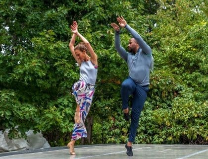 A man and woman doing a form of dance with their arms raised outdoors in front of full green trees at Windhover Performing Arts Center.