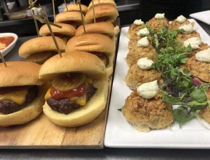 A close-up display of sliders on a wooden board and crabcakes with green garnish to the right of the sliders